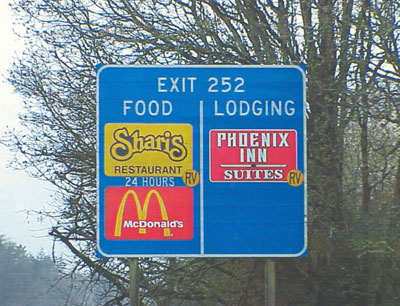 Highway sign showing the use of the RV Friendly symbol on a food-lodging sign.  The symbol is placed directly in the corner of the emblem it modifies.