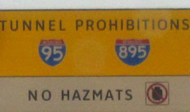 At lower left is a rectangular regulatory sign that displays the legend "TUNNEL PROHIBITIONS - NO HAZMATS" in the alternative alphabet in a negative-contrast orientation.