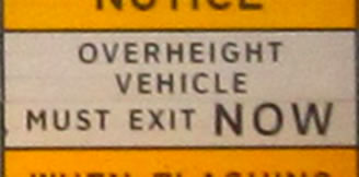 Lower right is a rectangular regulatory sign that displays the legend "OVERHEIGHT VEHICLES MUST EXIT NOW" in the alternative alphabet in a negative-contrast orientation.