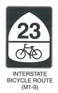 Pedestrian and Bicycle Sign "INTERSTATE BICYCLE ROUTE (M1-9)" is shown as a vertical rectangular black sign with a black legend. It shows an inverted white shield, with the numerals "23" in black at the top. Under the numerals, a horizontal black line is shown above a black symbol of a bicycle.