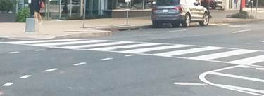 These two photos both show a crosswalk that is marked not only by a pair of white transverse lines that define the outside edges of the crosswalk, but also by a series of wide white longitudinal lines parallel to traffic flow between the transverse lines to make the crosswalk more visible to motorists.