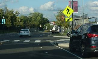This photo shows a motor vehicle lane and a bike lane just to the left of a parked motor vehicle.  The curb and sidewalk area on the right-hand side of the roadway extends out in front of the parked vehicle to the edge of the bike lane.  A marked crosswalk is shown across the roadway at the point where the curb and sidewalk area on the right-hand side is extended.  A Pedestrian (W11-2) warning sign with a diagonal downward pointing arrow (W16-7P) plaque is post-mounted on the right-hand side of the roadway adjacent to the crosswalk.
