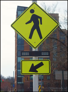 Figure 1a - pedestrian (W11-2) sign with a downward diagonal arrow (W16-7P) supplemental plaque mounted below it. A rectangular rapid flashing beacon light bar is mounted between the sign and the plaque. Both horizontally-arranged yellow rectangular indications on the light bar are dark.