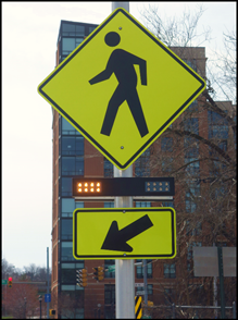 Figure 1b - pedestrian (W11-2) sign with a downward diagonal arrow (W16-7P) supplemental plaque mounted below it.  A rectangular rapid flashing beacon light bar is mounted between the sign and the plaque.  The yellow rectangular indication on the left-hand side of the light bar is illuminated and the yellow rectangular indication on the right-hand side of the light bar is dark.
