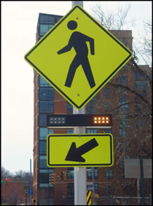 Figure 1c - pedestrian (W11-2) sign with a downward diagonal arrow (W16-7P) supplemental plaque mounted below it. A rectangular rapid flashing beacon light bar is mounted between the sign and the plaque. The yellow rectangular indication on the left-hand side of the light bar is dark and the yellow rectangular indication on the right-hand side of the light bar is illuminated.