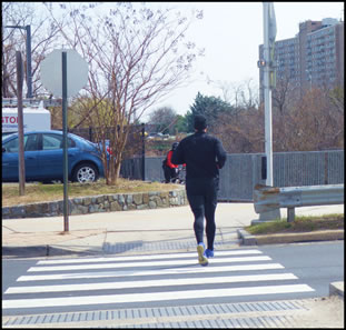 Figure 2a - a small white pilot light that is mounted on the side of a small box that is mounted on a pole at a height of approximately 8 feet on the far side of the street. The pilot light is facing pedestrians who are crossing the roadway in the crosswalk. The photograph is a view from a distance showing a pedestrian crossing in the crosswalk, and the pilot light, box, and support pole can be seen at the far end of the crosswalk.