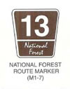 Guide Sign "NATIONAL FOREST ROUTE MARKER (M1-7)" is shown as a brown trapezoid with a slightly wider top and a white border and legend. The large white numerals "13" are shown in the top two-thirds of the sign above a horizontal white line that extends across the width of the sign. Below the line, the words "National Forest" are shown in white cursive lettering on two lines at the bottom of the sign. The sign is labeled "Forest Route Sign."