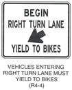 Pedestrian and Bicycle Sign "VEHICLES ENTERING RIGHT TURN LANE MUST YIELD TO BIKES (R4-4)" is shown as a horizontal rectangular white sign with a black border and legend. It shows the words "BEGIN RIGHT TURN LANE" on two lines above a diagonal arrow pointing down and to the left above the words "YIELD TO BIKES."